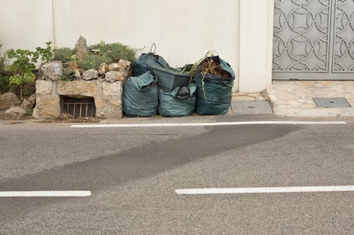 The image shows three large, dark green refuse bags filled with mixed waste, placed on the edge of a paved street near a white wall with a small garden feature to the left, consisting of rocks and green plants. The refuse bags are situated close to a building wall and partially obstruct a metal drainage grate embedded in the pavement. The street has white line markings along the edge and appears clean, with no other visible litter or debris in the surrounding area. The environment suggests a location within a residential or urban area, possibly in Ware, where Commercial Waste Ware offers rubbish removal services. The scene emphasizes waste collection materials ready for disposal or clearance, reflecting typical rubbish separation at the curbside outside a property.