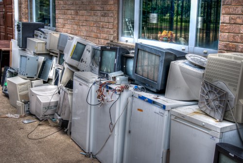 A row of discarded, outdated computer monitors and desktop towers stacked along a brick building exterior, positioned on a concrete pavement. The monitors vary in size and style, with some having curved screens and others flat, displaying worn or blank screens, and many are linked by tangled cables and wires. The desktop units and monitors show signs of age, with some casing tarnished or stained, and the arrangement appears haphazard, with items leaning or resting against each other. Behind the setup, a window with reflective glass reveals some greenery outside, and the surrounding environment suggests no organized disposal, emphasizing the need for professional rubbish removal services. Commercial Waste Ware would typically handle such electronic waste collection to ensure responsible disposal in line with environmental standards, especially in a town within the Ware postcode area. The overall scene is well-lit, capturing the textures of the aged plastic and metal components and providing a clear view of the electronic waste cluttering the exterior space.