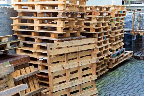 A large stack of wooden pallets arranged in a neat, overlapping formation, situated outdoors on a paved area adjacent to a stone wall and metal fencing. The pallets are light-colored, made of smooth, untreated timber with slatted surfaces and supporting beams, showing slight variations in size and condition. Some pallets have visible nail heads and minor wear. The stack appears to be part of a commercial waste storage area, likely associated with waste removal services in Ware. There are additional pallets and wooden items visible in the foreground, and the background reveals a partially obscured industrial or storage environment with fencing and a building. The lighting suggests a cloudy day, creating soft, diffuse illumination that highlights the natural wood textures and the urban outdoor setting. Overall, the scene reflects an organized accumulation of pallets ready for recycling or waste collection, relevant to local rubbish removal services operated by Commercial Waste Ware.