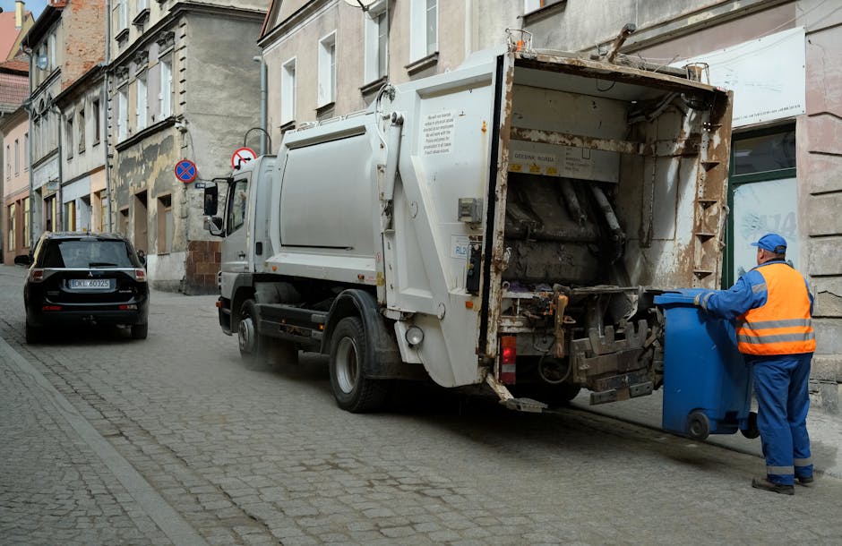 A large accumulation of various types of waste and rubbish is seen piled up next to a sidewalk in an urban environment. The refuse includes black plastic bags, flattened cardboard boxes, paper and car