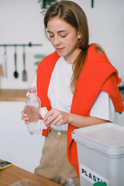 A young woman with long brown hair, wearing a white T-shirt and a red vest, stands at a kitchen counter sorting waste for recycling. She is holding a crumpled brown paper bag, examining it carefully.